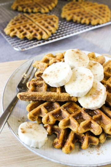 Stacked pumpkin waffles topped with banana slices and sprinkles of cinnamon and drizzled with syrup