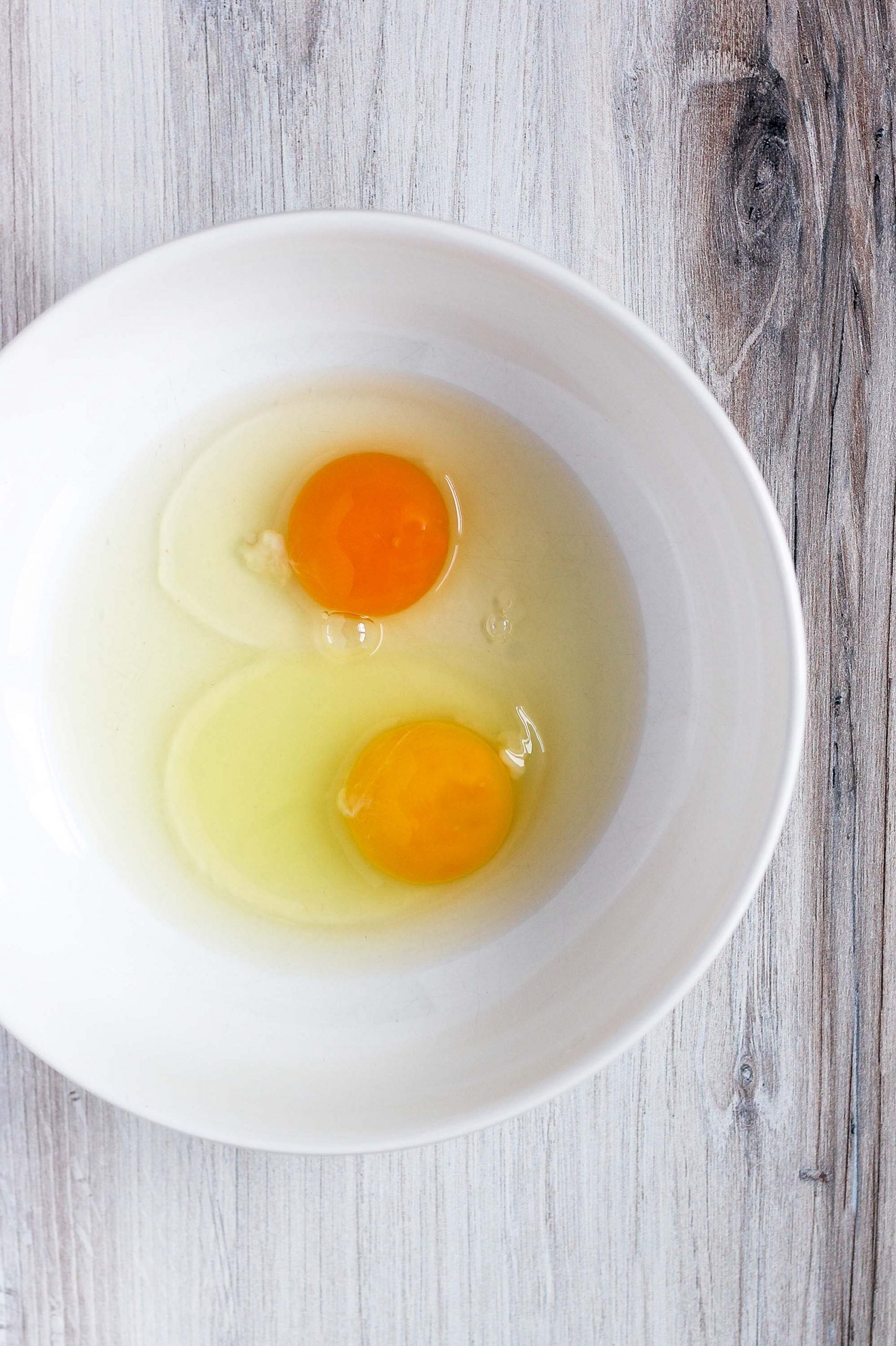 2 cracked eggs in a white bowl - one yolk is brighter orange