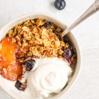 overhead view of peach and blueberry crisp in white bowl topped with coconut whipped cream