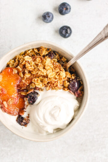 overhead view of peach and blueberry crisp in white bowl topped with coconut whipped cream