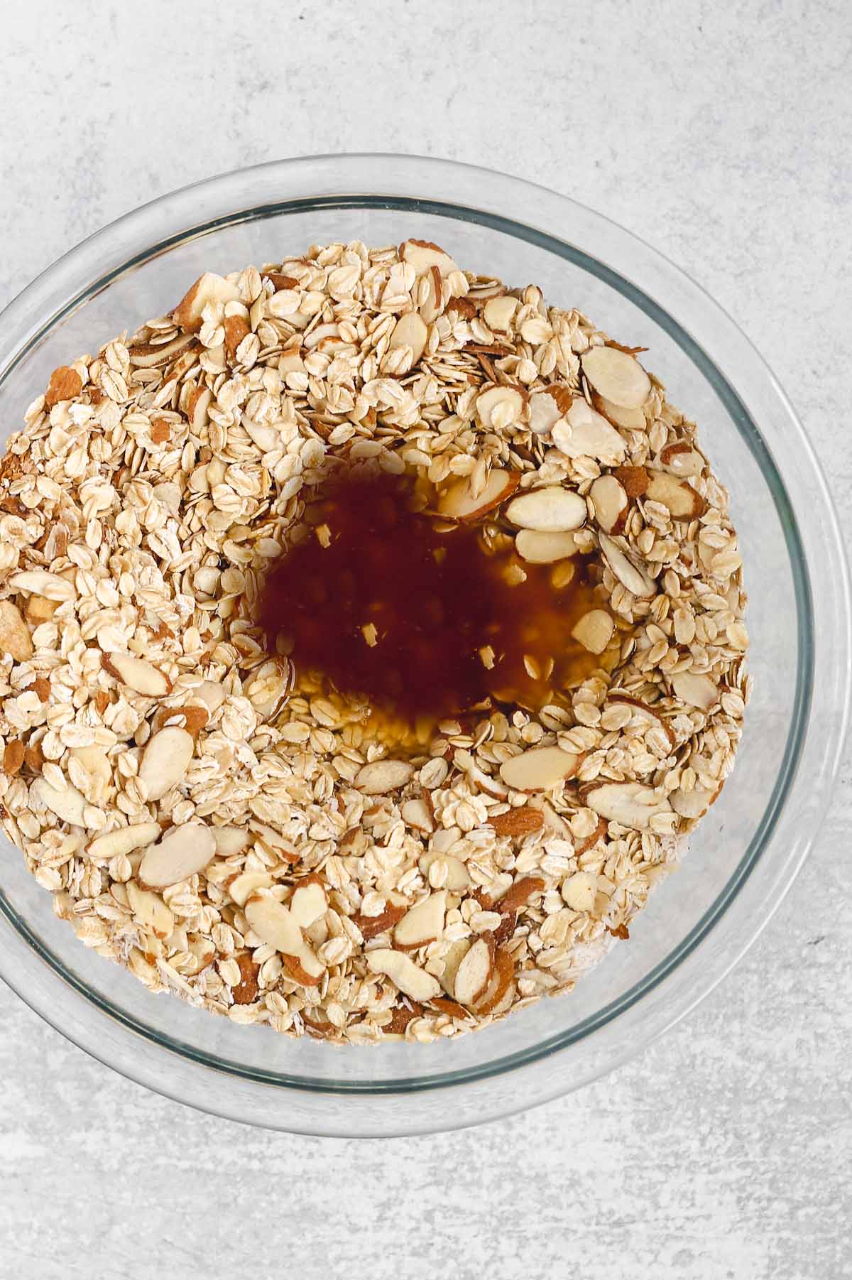 Dry ingredient needed for homemade granola in a glass mixing bowl with wet ingredients poured on top.