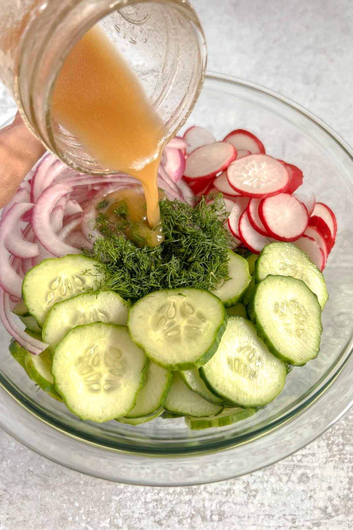 Vinaigrette being poured over cucumber radish salad ingredients in a glass mixing bowl.