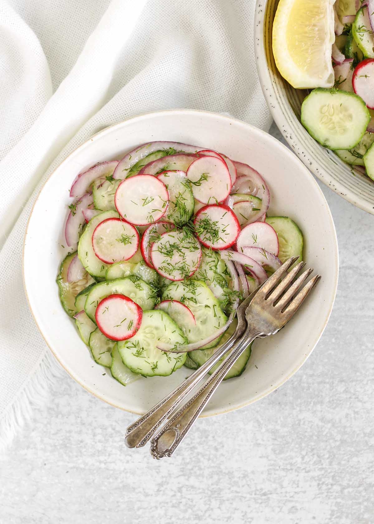 Cucumber radish salad on a small plate with two vintage forks and a bowl of the salad off to the side.