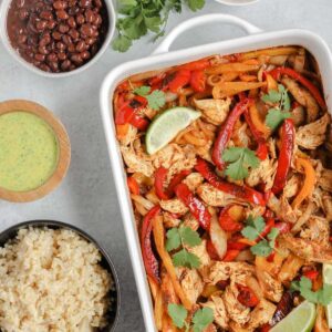 baked chicken and peppers in a white baking dish with a bowl or rice, beans, and dressing on the side