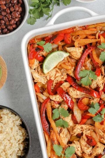 baked chicken and peppers in a white baking dish with a bowl or rice, beans, and dressing on the side
