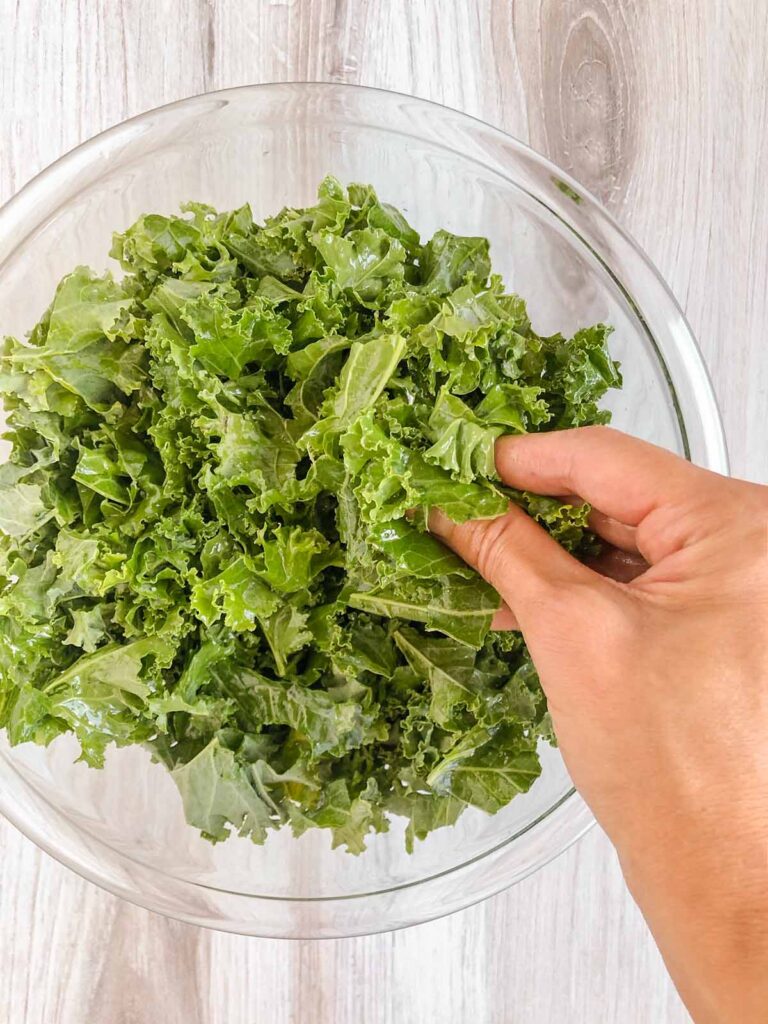 A hand massaging kale in a glass mixing bowl.