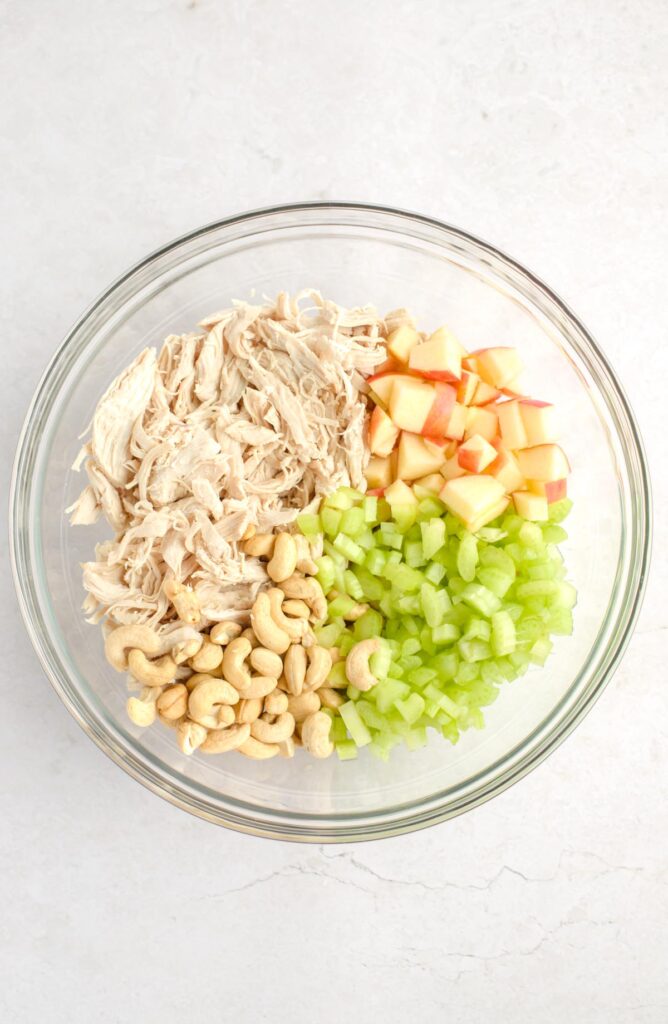 Shredded chicken, diced apples, diced celery, and cashews in a mixing bowl.