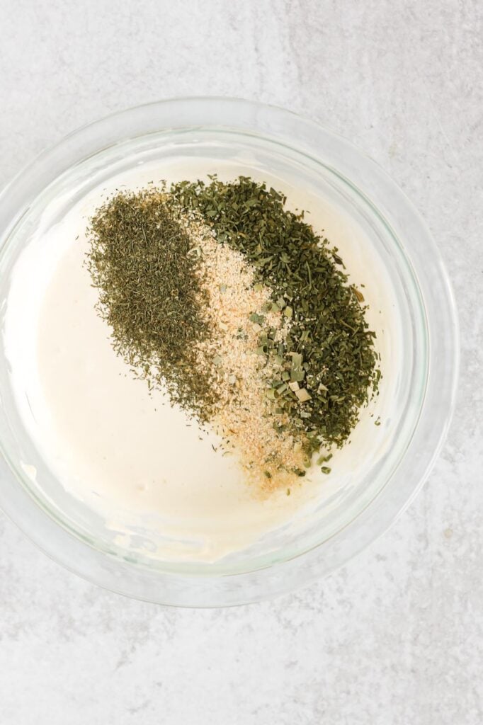 Yogurt and mayo mixed together in a glass mixing bowl with dried herbs added to the bowl.