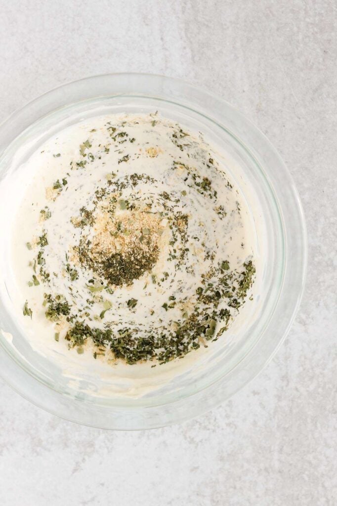 Mayonnaise, yogurt, and herbs being mixed in a mixing bowl.
