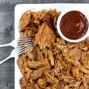 Pulled pork with two forks and a side of BBQ sauce on a plate.