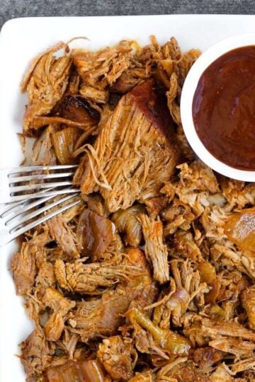 Pulled pork with two forks and a side of BBQ sauce on a plate.