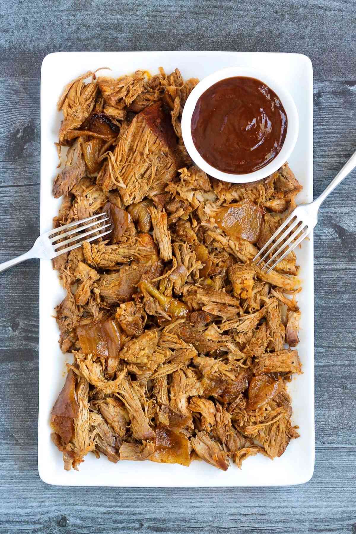 Pulled pork and a side of BBQ sauce with two forks on a serving tray.
