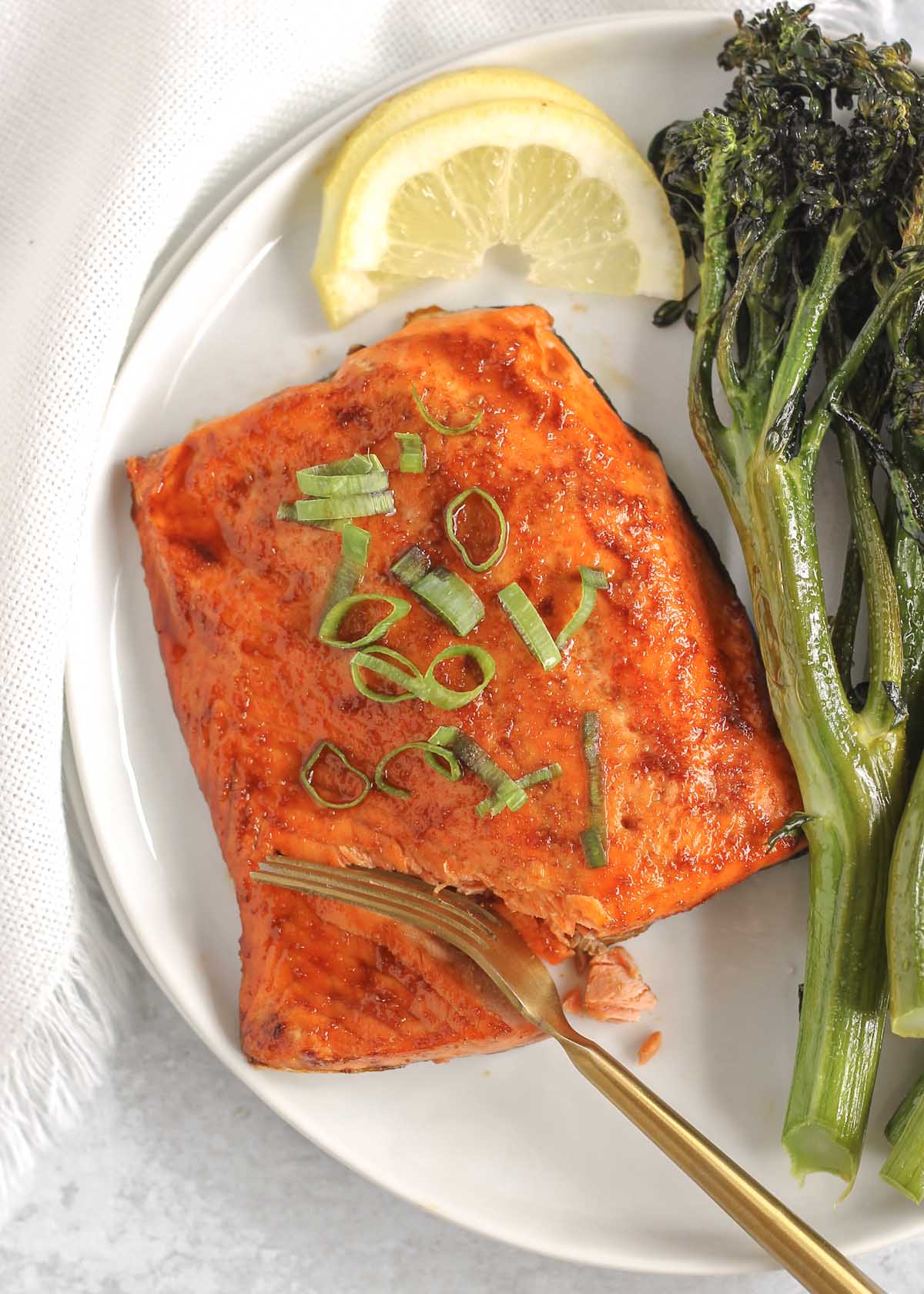 A fork cutting into a salmon fillet on a plate with a side of broccolini and lemon.