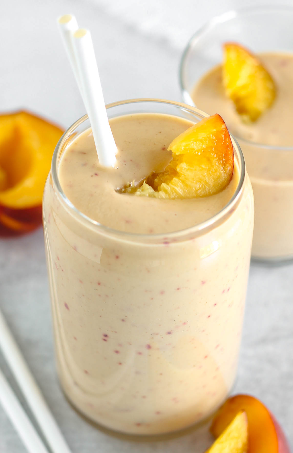 Tall glass filled with a peach banana smoothie topped with a peach slice and two straws.