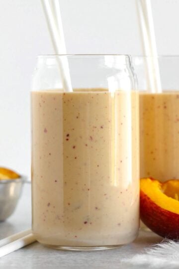 Two glasses filled with peach smoothie with half a peach laying next to the glasses.