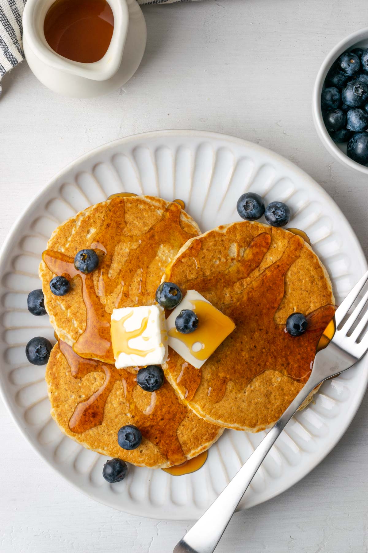 Three oat flour pancakes topped with butter, blueberries, and syrup on a round white plate.