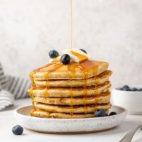 Syrup being poured over a stack of oat flour pancakes topped with blueberries.