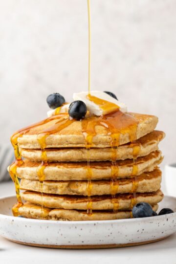 Syrup being poured over a stack of oat flour pancakes topped with blueberries.