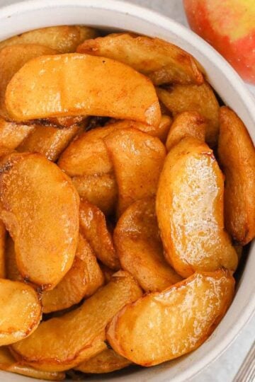 Air fried apples sprinkled with cinnamon in a small white bowl with a spoon next to the bowl.
