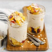 Two mason jars on a wood cutting board filled with overnight oats topped with peaches, almonds, and coconut. Oats sprinkled on the cutting board.