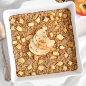 Baked apple oatmeal topped with apple slices in a square white baking dish with apples and syrup on the side.