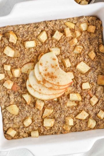 Baked apple oatmeal topped with apple slices in a square white baking dish with apples and syrup on the side.