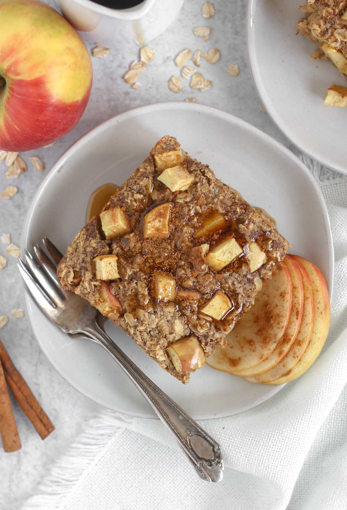 Square piece of apple cinnamon baked oatmeal topped maple syrup on a round plate with a vintage fork.
