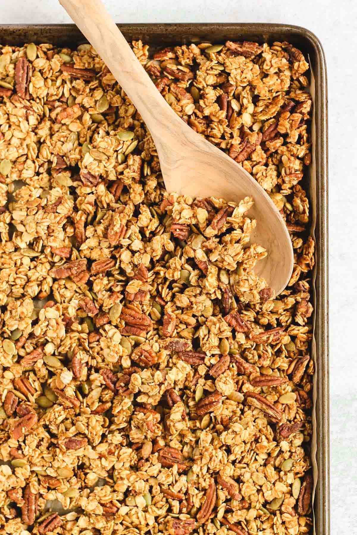 Pumpkin granola on a large baking sheet with a wooden spoon scooping up some granola.