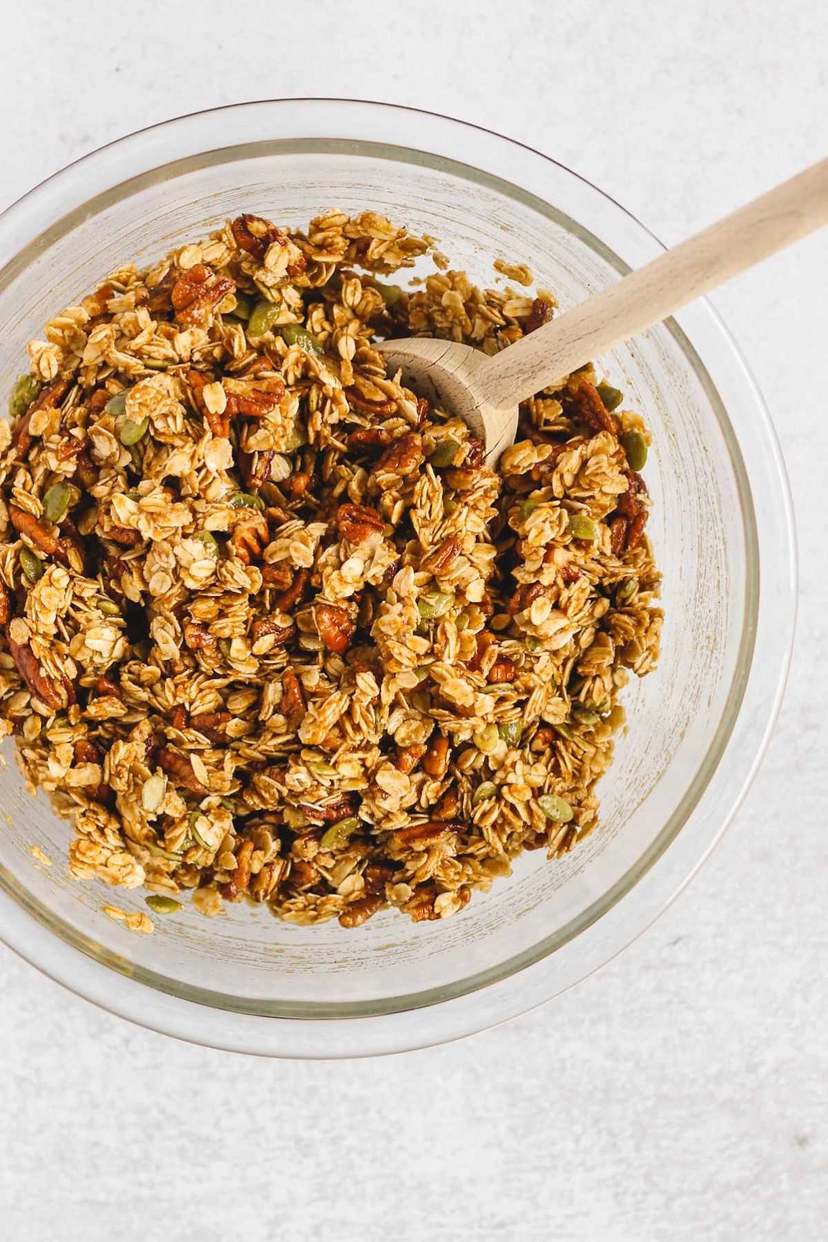 Pumpkin granola ingredients mixed together in a glass mixing bowl with a wooden spoon.