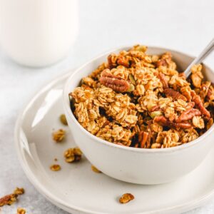 Pumpkin granola in a bowl on a small plate with a glass jar of milk behind bowl.