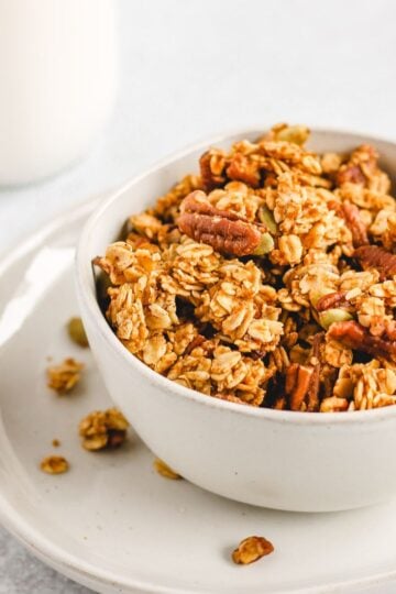 Pumpkin granola in a bowl on a small plate with a glass jar of milk behind bowl.