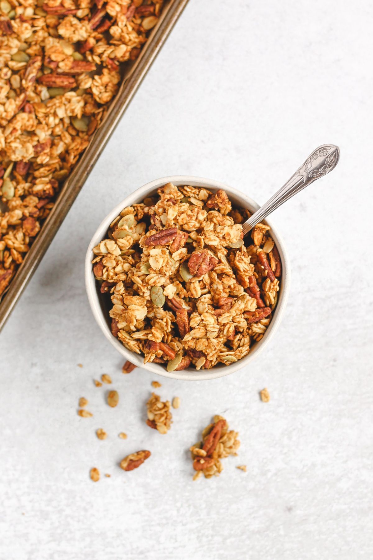 Pumpkin granola in a small bowl with a vintage spoon sitting next to baking sheet with granola.