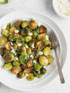 Roasted Brussels sprouts with goat cheese on a round plate with a vintage fork with raw Brussels in the background.