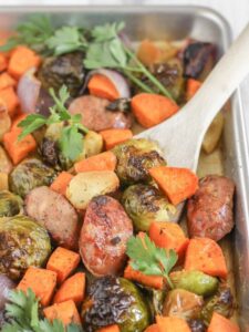 Roasted vegetables and chicken sausage on a large baking sheet with a wooden spatula scooping up some of the food.