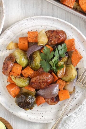 Chicken sausage with roasted veggies on a round white plate topped with parsley with a vintage fork on the plate.