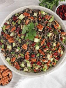 Wild rice salad with pecans, dried cranberries, and apples in a ceramic serving bowl with a vintage serving spoon in the salad.
