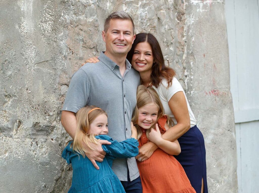 Mom, dad, and two daughters standing next to a gray wall and hugging.