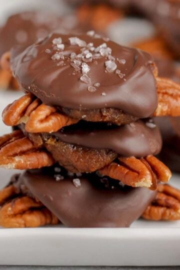 Chocolate turtle candies stacked 3high on a white serving tray and extra turtles in the background.