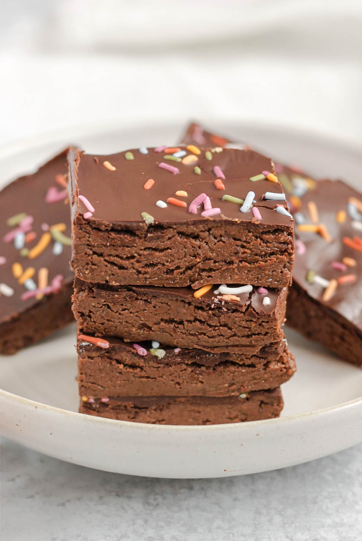 Square pieces of chocolate protein bar stacked 4 high on a plate with other bars in the background.