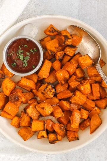 Air fried sweet potato cubes with a vintage serving spoon and side of ketchup on a round cream plate.
