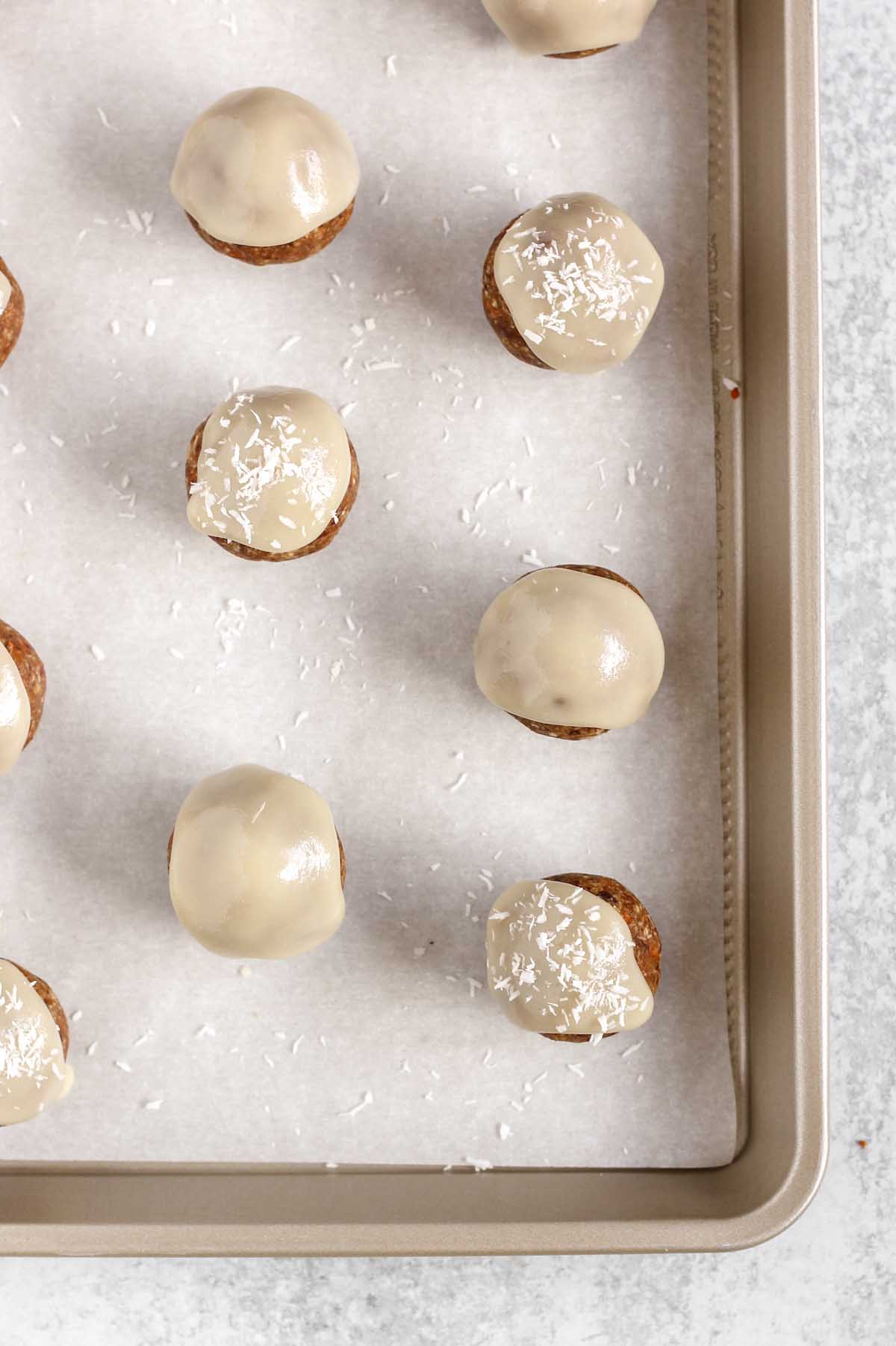 Carrot cake energy balls topped with glaze and sprinkled with coconut flakes on a cookie sheet.