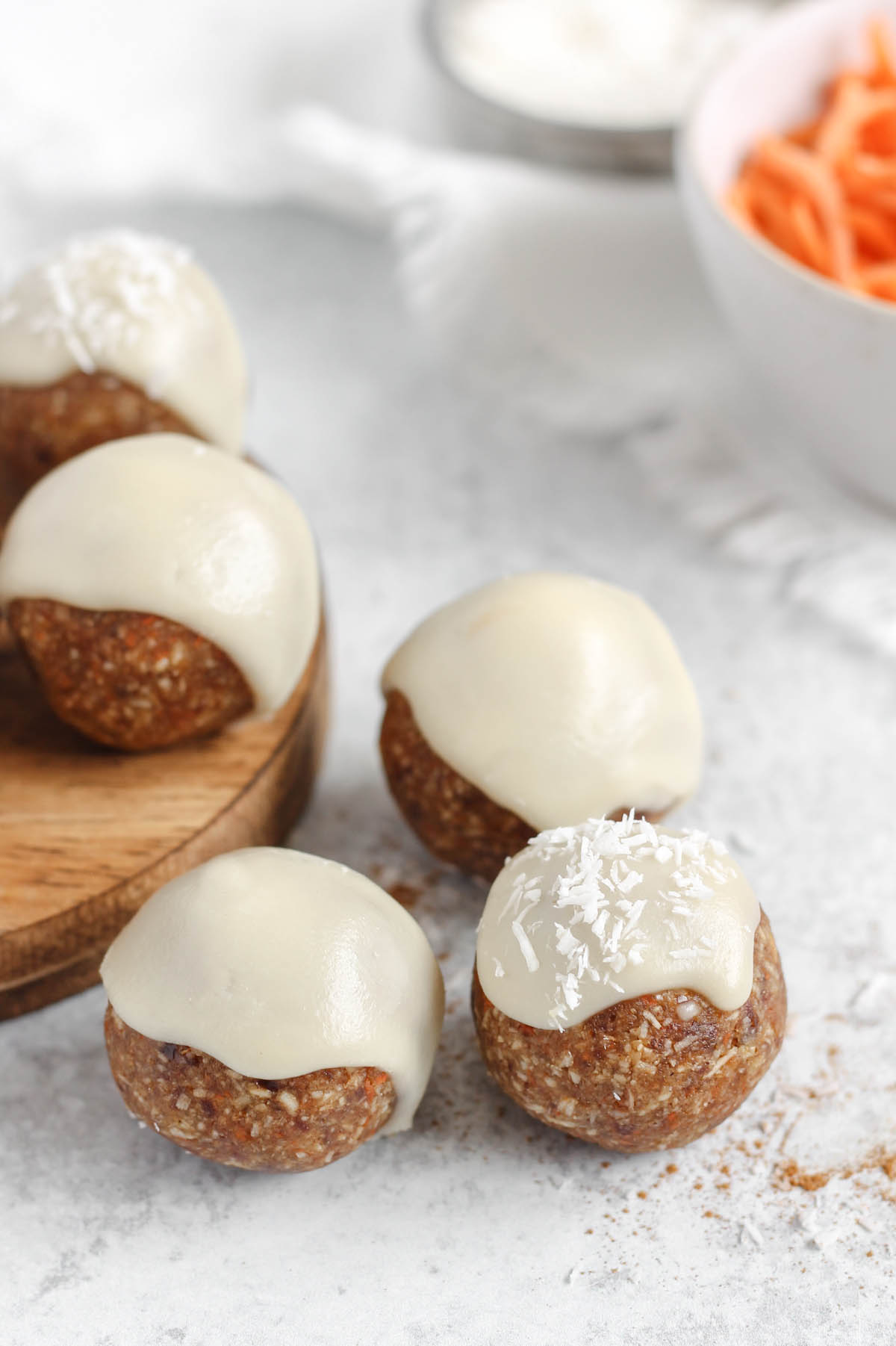 Three carrot cake bites topped with glaze on a gray surface with a bowl of shredded carrots in the background.