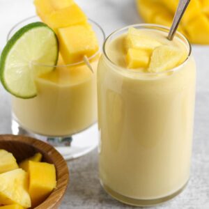Two glasses filled with a creamy yellow smoothie topped with fresh pineapple and mangos.