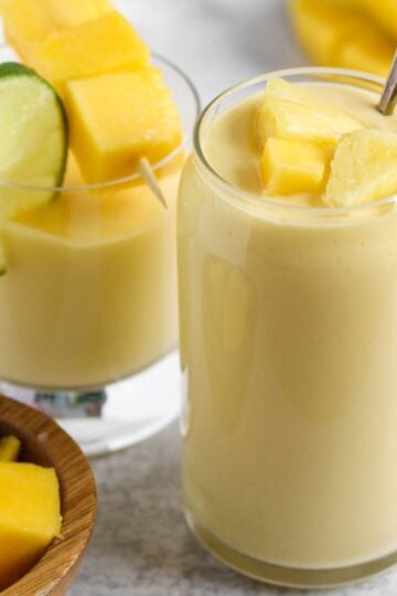 Two glasses filled with a creamy yellow smoothie topped with fresh pineapple and mangos.