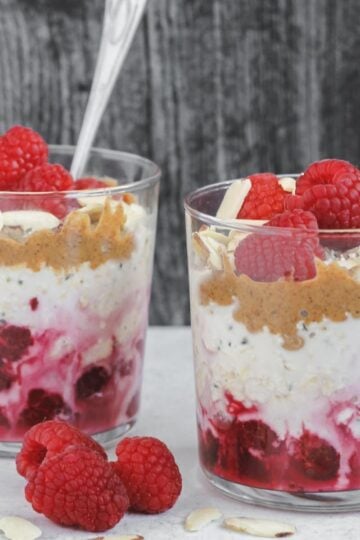 Two glass cups filled with layers of oats, raspberries, almond butter and almonds.