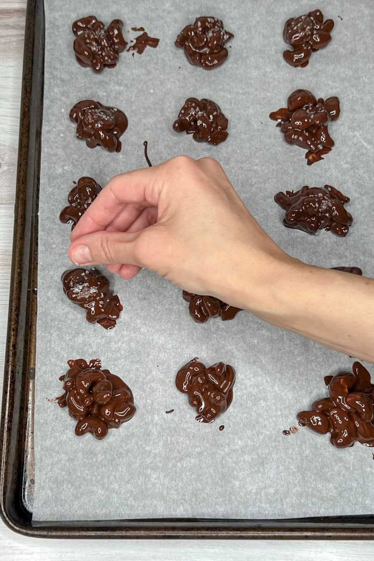 Hand sprinkling sea salt over chocolate cashew clusters on a baking sheet.