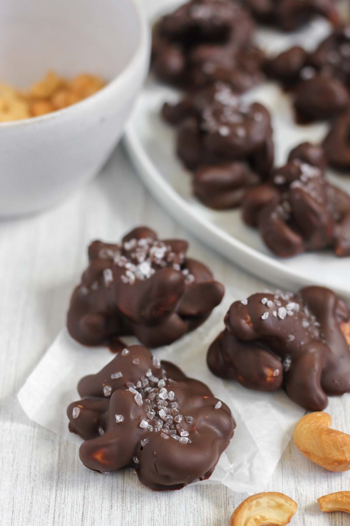 Three chocolate covered cashew clusters topped with sea salt with more cashew clusters in the background.