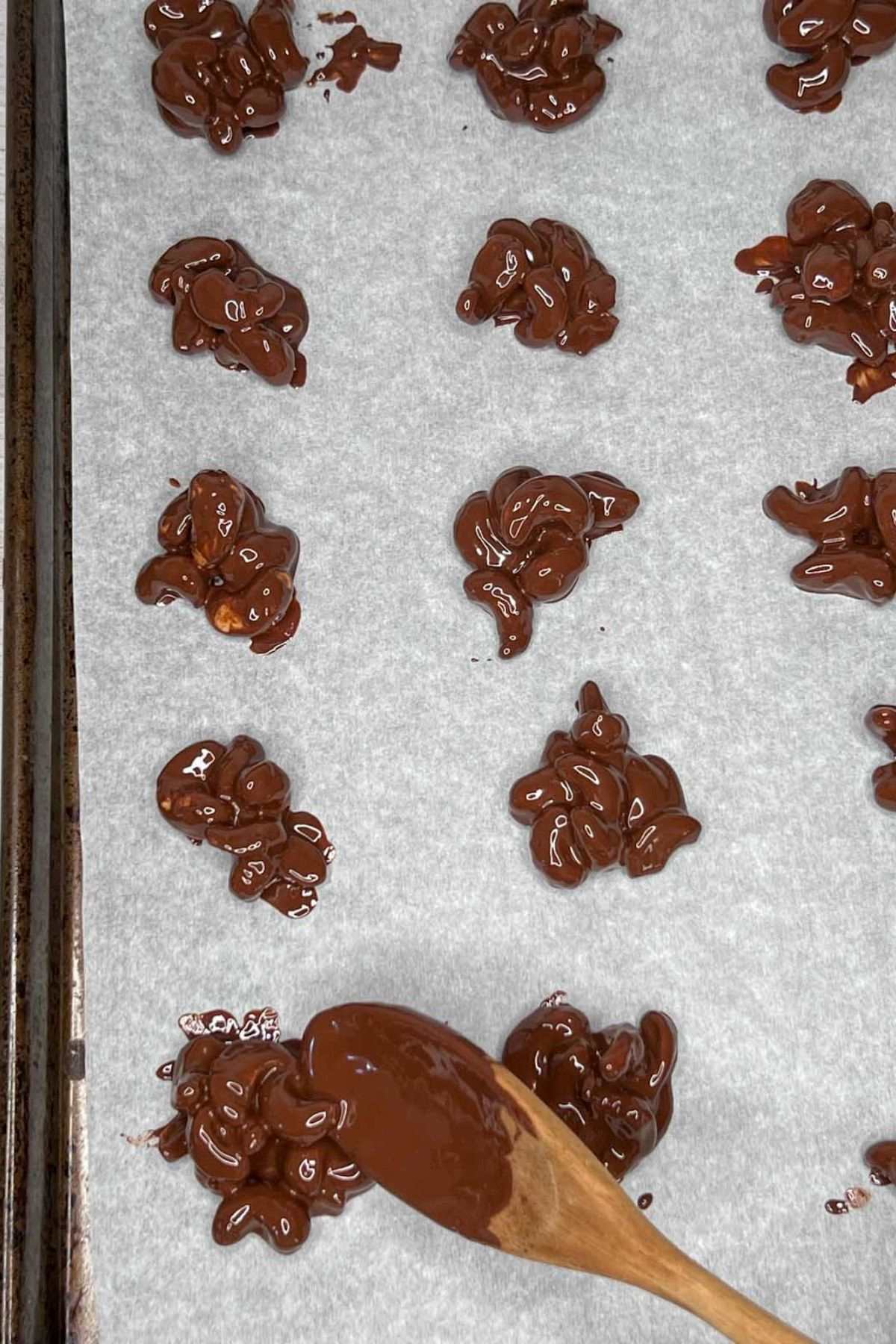 Wooding spoon dropping clusters of cashews covered in melted chocolate on a parchment paper lined baking sheet.