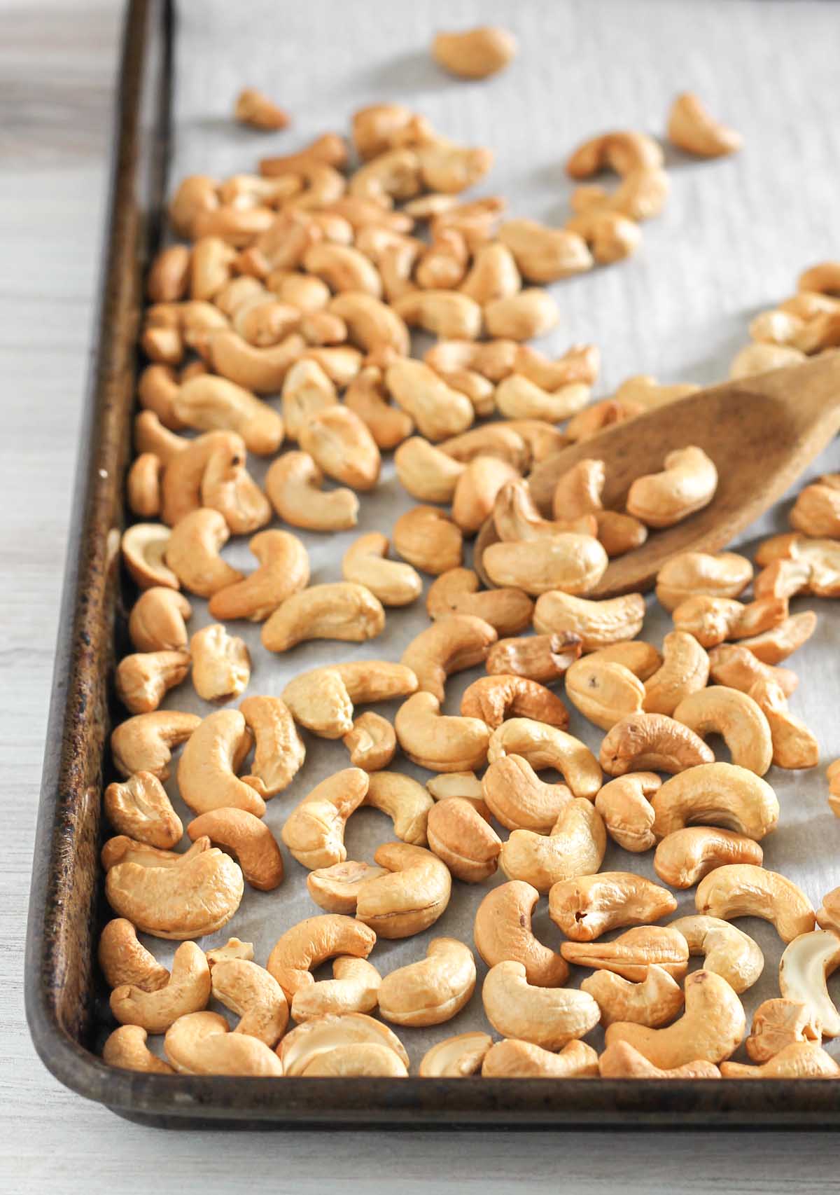 Wooden spoon scooping up roasted cashews on a vintage baking sheet.