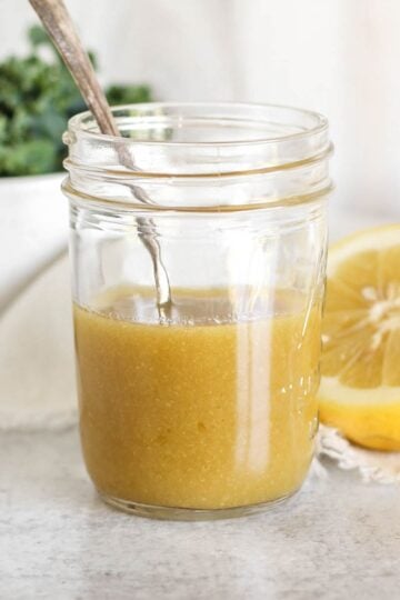 Maple Dijon dressing and a spoon in mason jar with a bowl of greens and a sliced lemon on the background.